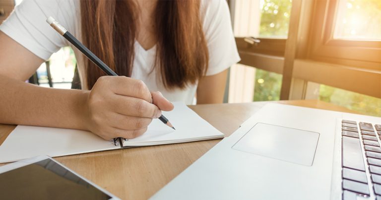 Woman writing in notebook while on laptop