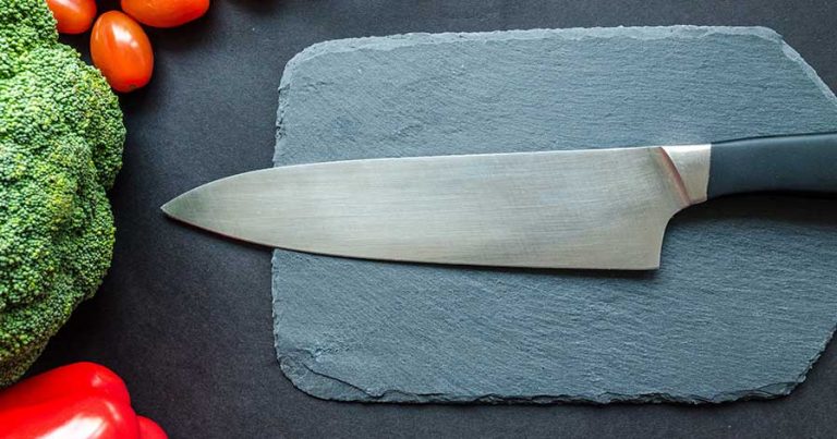 A knife on a cutting board, sitting beside vegetables