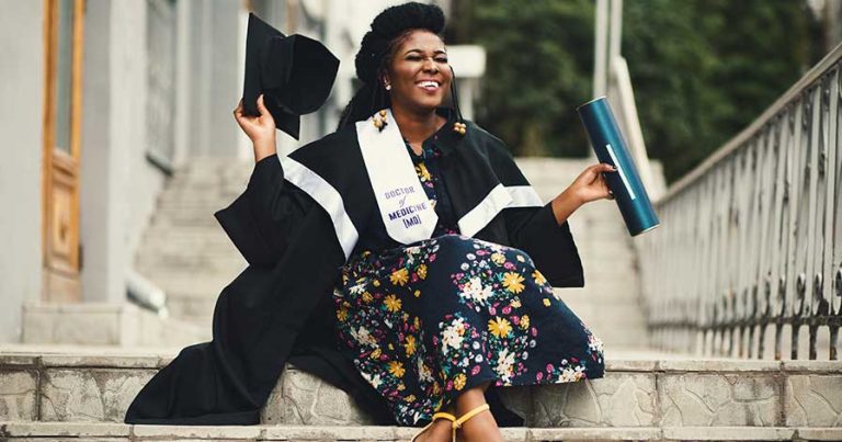 A woman sitting on steps, holding her college degree after a graduation ceremony