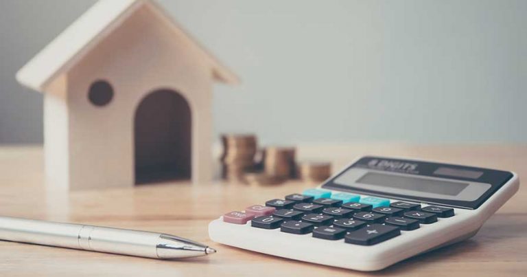 A calculator sitting on a table with a pen, and a wooden house in the background