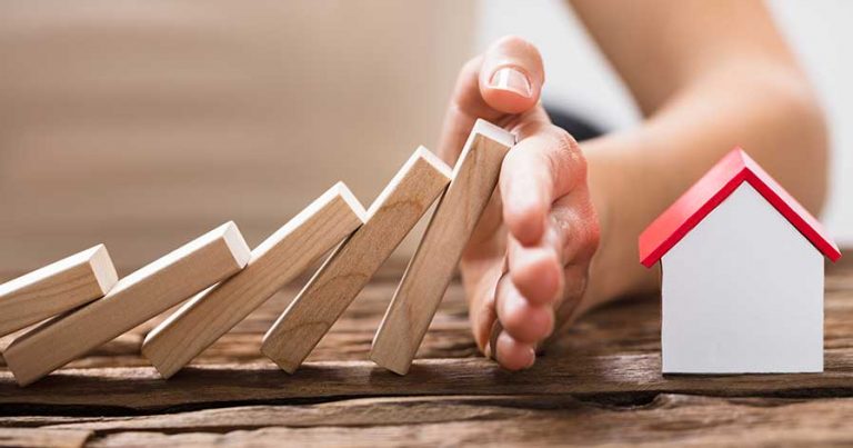 Dominos falling towards a toy home, with a hand stopping them