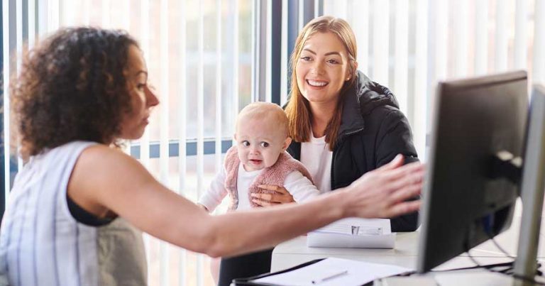 A government worker pointing to a computer screen while sitting with a mom and her baby.