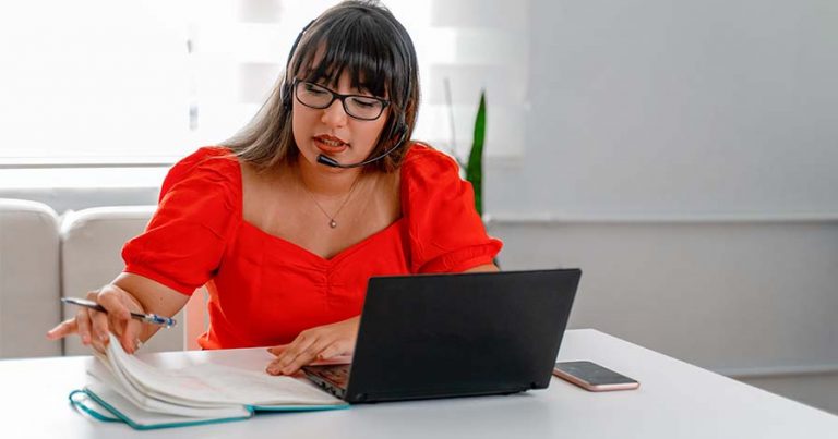 A woman sitting at a white table in front of an open laptop and notebook wearing a headset. a heads