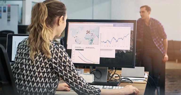 A lady working on a desktop computer that has graphs on the screen.