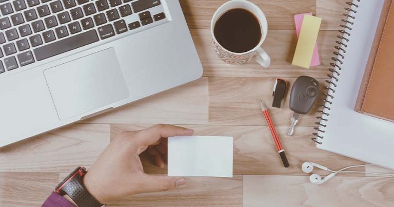 A flat lay of a person holding a white business card surrounded by a laptop keyboard, a mug full of coffee, a pen, car keys and a notebook.