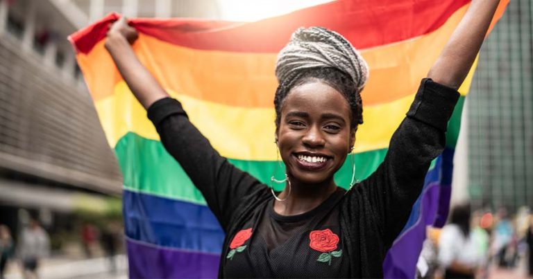 A woman standing on a street holding a pride flag behind her.