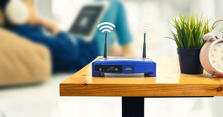 A blue internet router on a brown wooden table next to a plant and a clock.