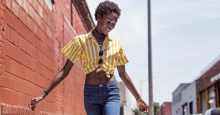 A woman outside wearing blue jeans and a yellow blouse crop top.