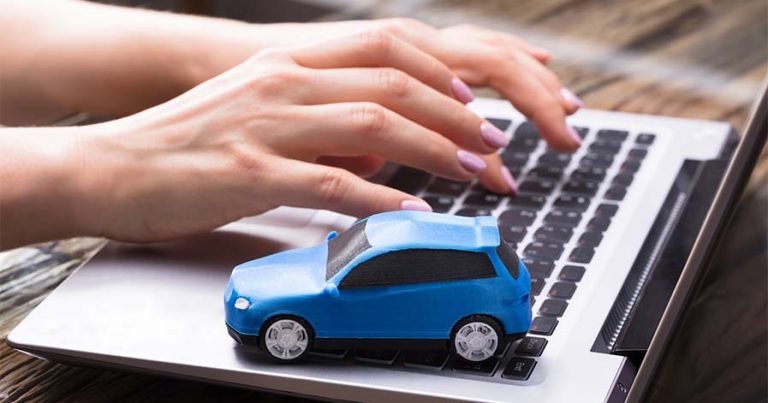 A small blue toy car on a laptop as someone types on the keyboard.