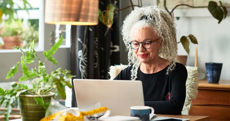 An older lady sitting in front of a laptop at a table with plants around her.