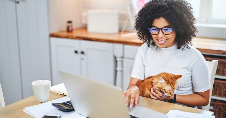 A woman sitting at a table in front of a laptop with an orange cat on her lap.