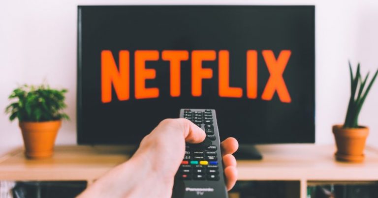 A hand holding a remote up in front of a TV, displaying "Netflix" on the screen.
