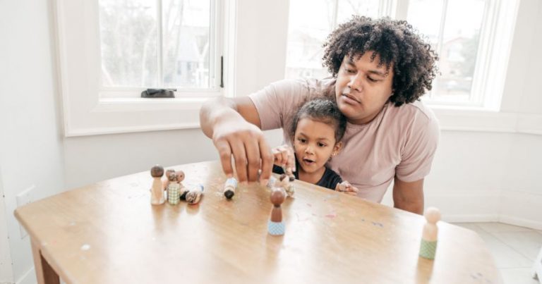 A man playing with a little girl at a table, holding dolls.