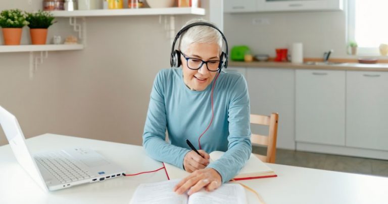An older woman with headphones on, writing in a notebook in front of a laptop